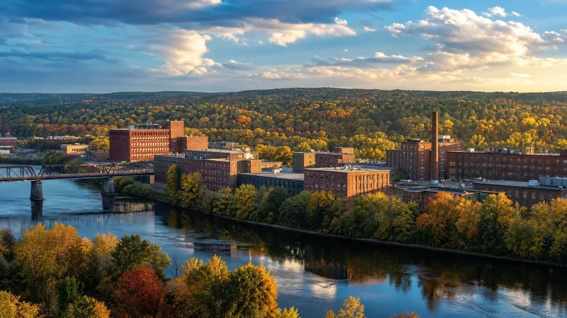 Inland river city downtown of brick mills and a tree-lined riverbank with pine forest beyond, early fall color, Bangor Maine Business License